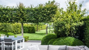An outdoor patio with a grey sofa, white table, and chairs, surrounded by greenery and a hedge. - La Cereza (Oudenaarde)