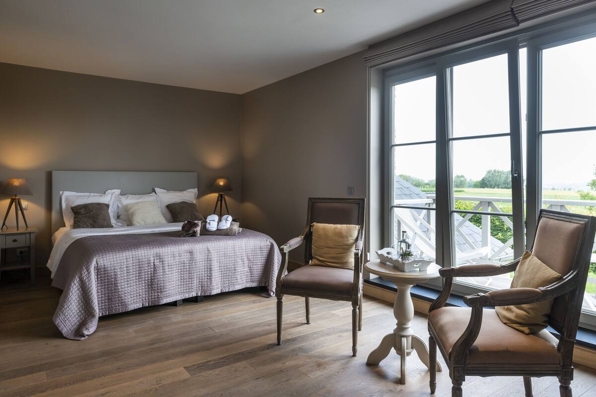 A bedroom with a large bed, two chairs, a small table, and a window with a view of the outdoors.