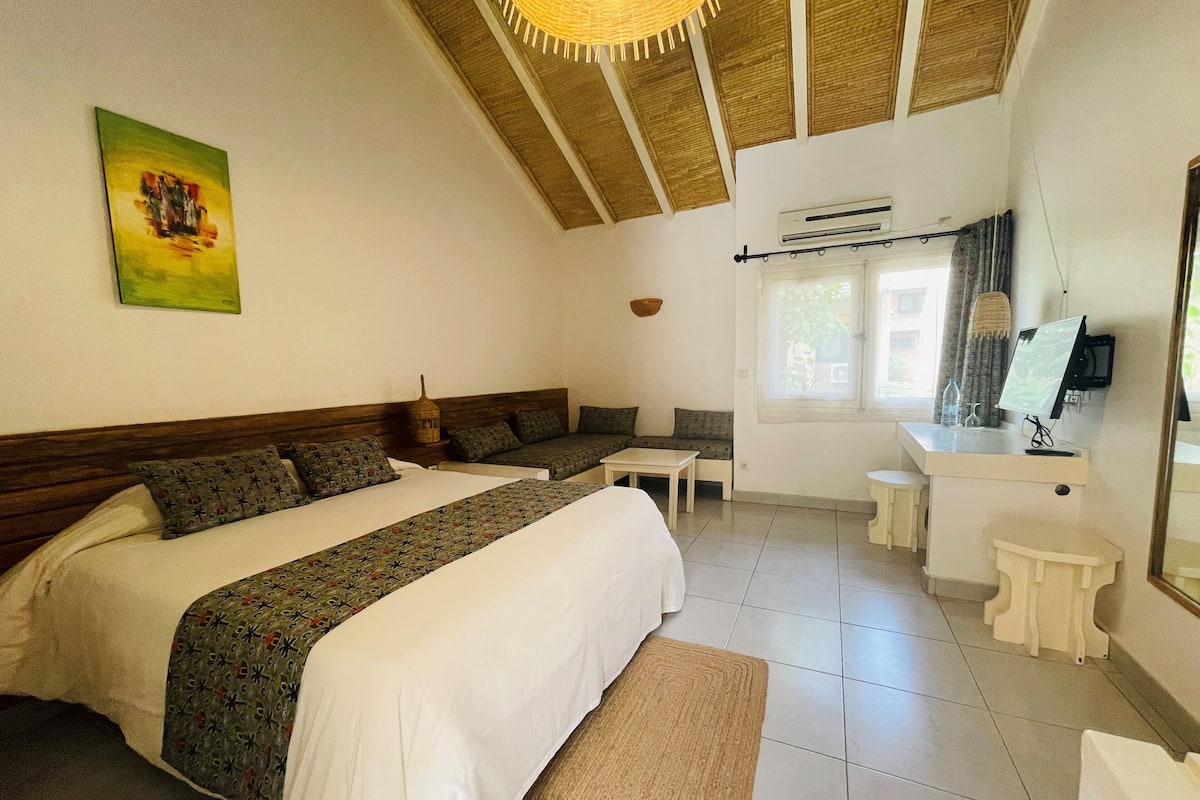 A hotel room with a large bed, a sofa, a small table, a mirror, and a window with curtains.