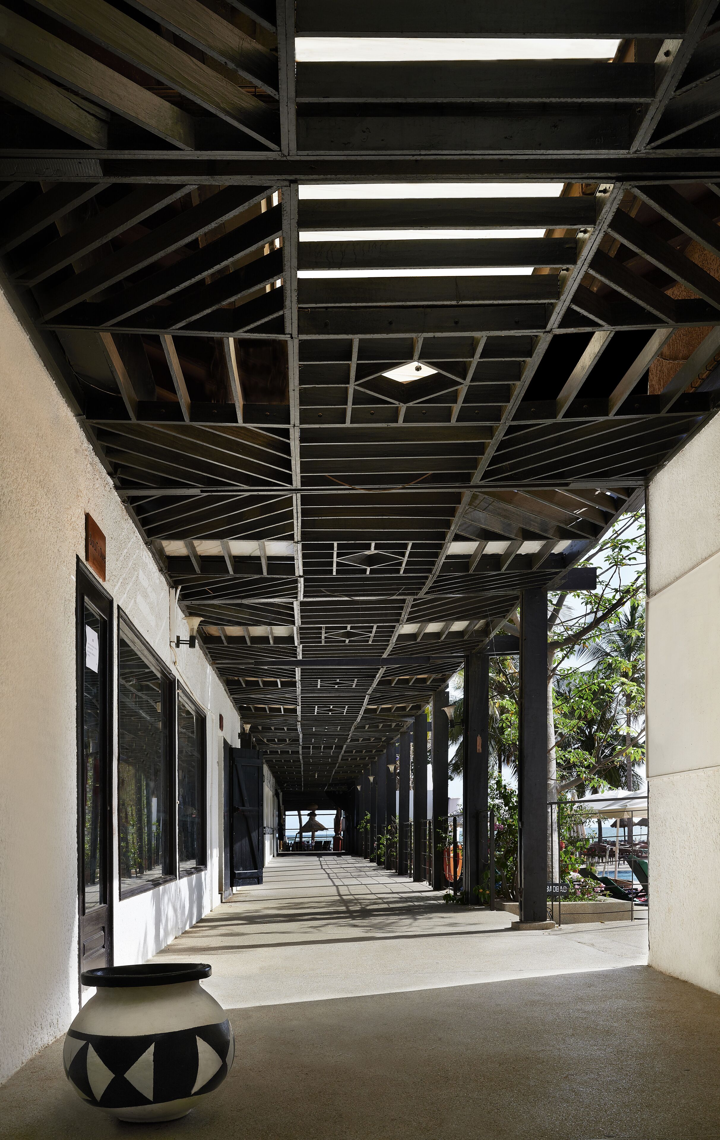 Un pasillo cubierto con estructura de techo metálico, ventanas grandes y una planta en maceta.