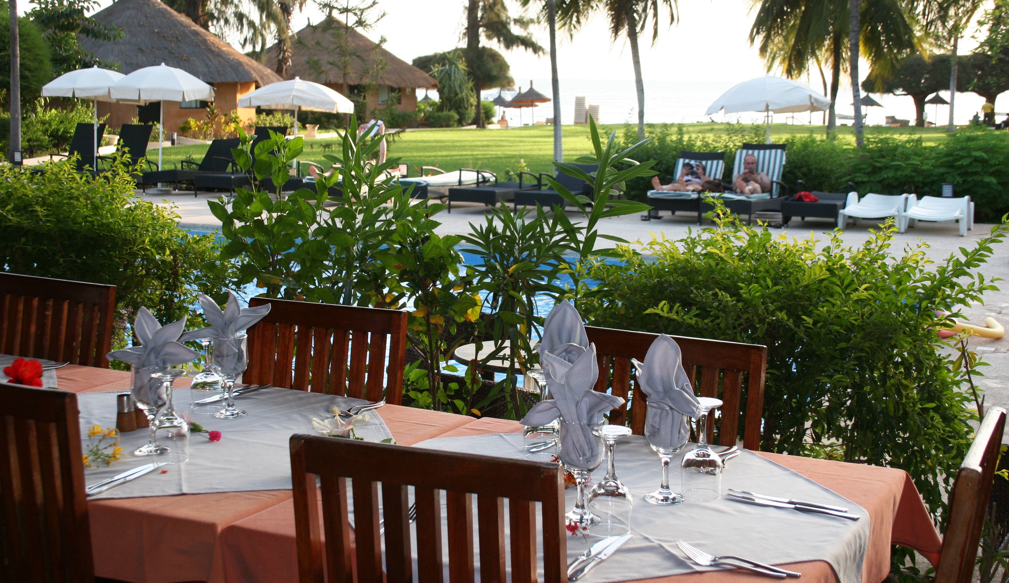 An outdoor dining area with tables set for two, surrounded by lush greenery and a view of the ocean.