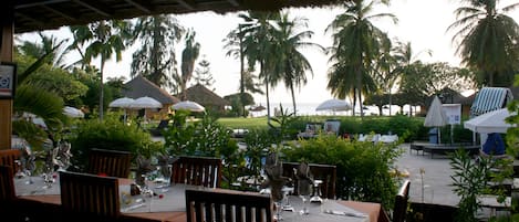 An outdoor restaurant with tables set for dining, surrounded by palm trees and thatched-roof umbrellas.