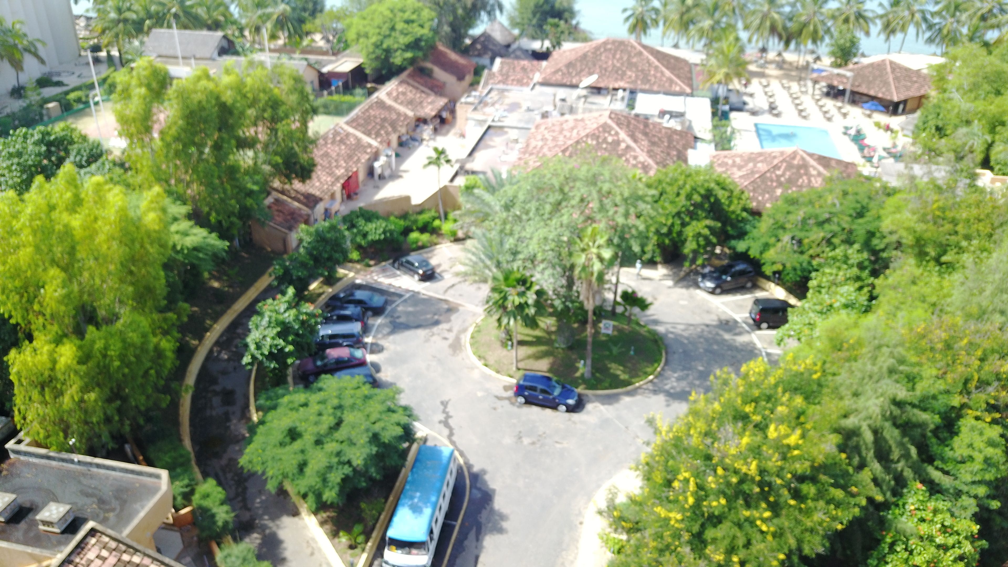 A parking area with several cars, a bus, and a roundabout. There are trees and buildings with red-tiled roofs.