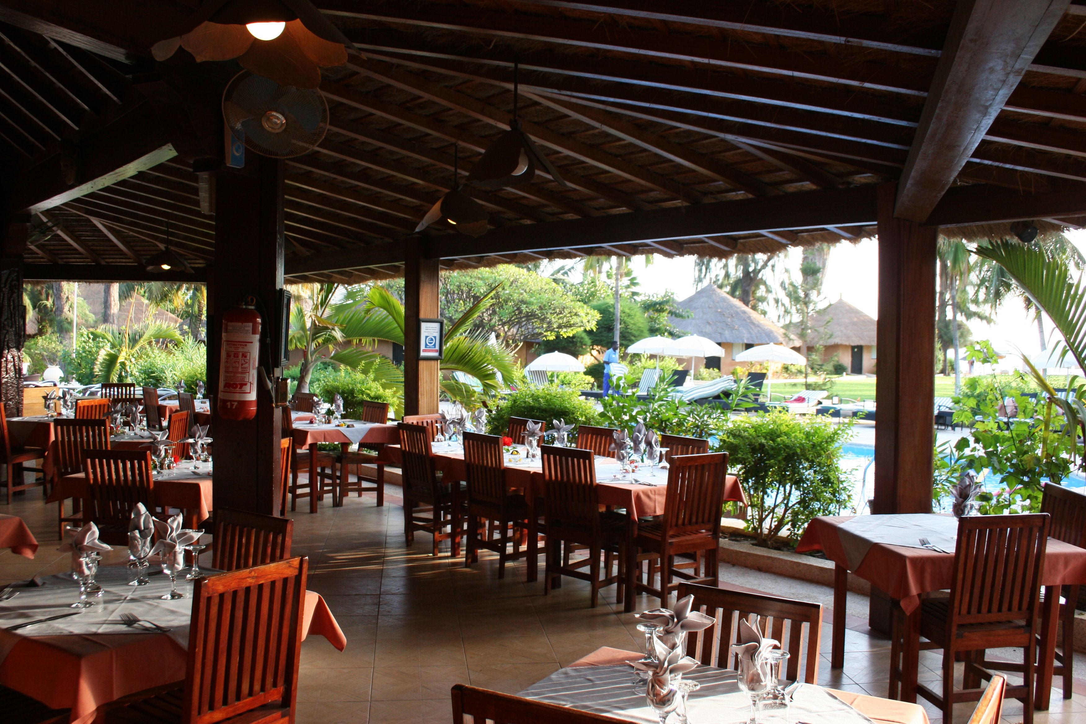 Un restaurante al aire libre con mesas y sillas de madera, piso de baldosas y vistas a una piscina y vegetación.
