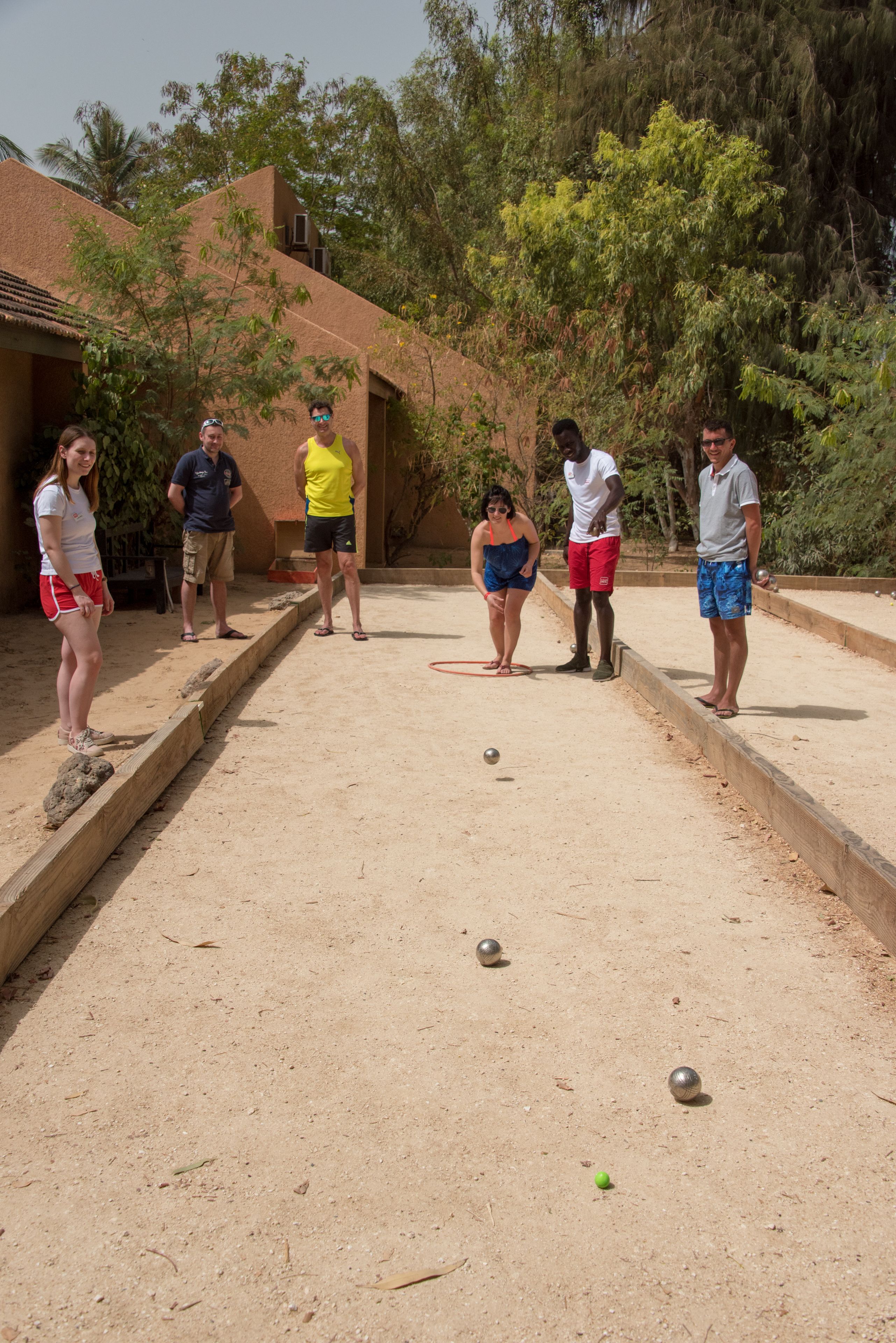 Un grupo de personas jugando al juego de bolos en un espacio al aire libre con suelo de arena y límites de madera.