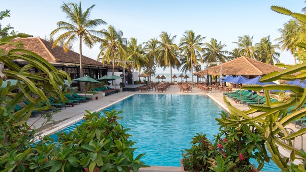 Área de piscina com espreguiçadeiras e guarda-sóis, cercada por palmeiras e um edifício com telhado de azulejos.