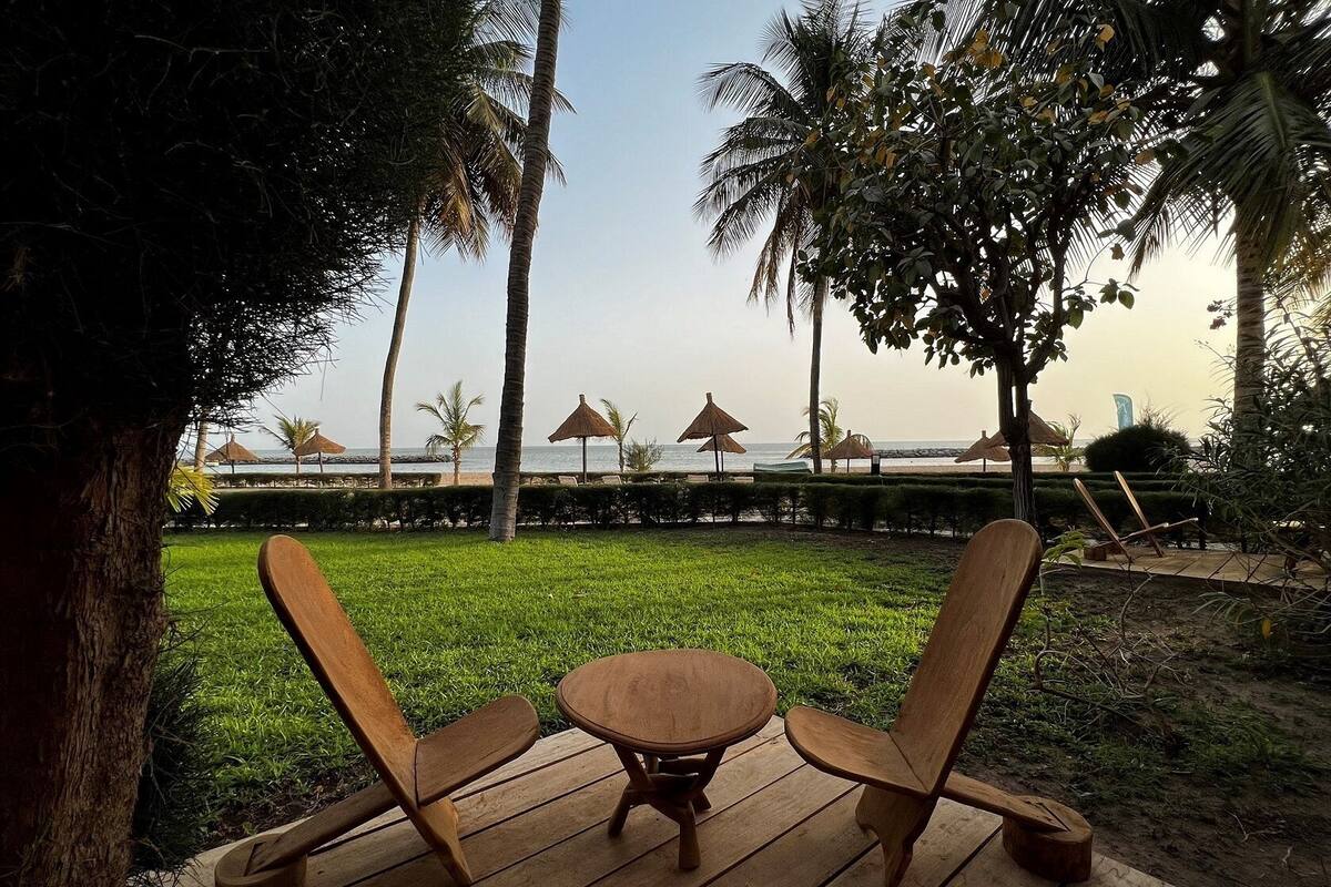 A wooden deck with a table and two chairs, overlooking a beach with palm trees and thatched umbrellas.