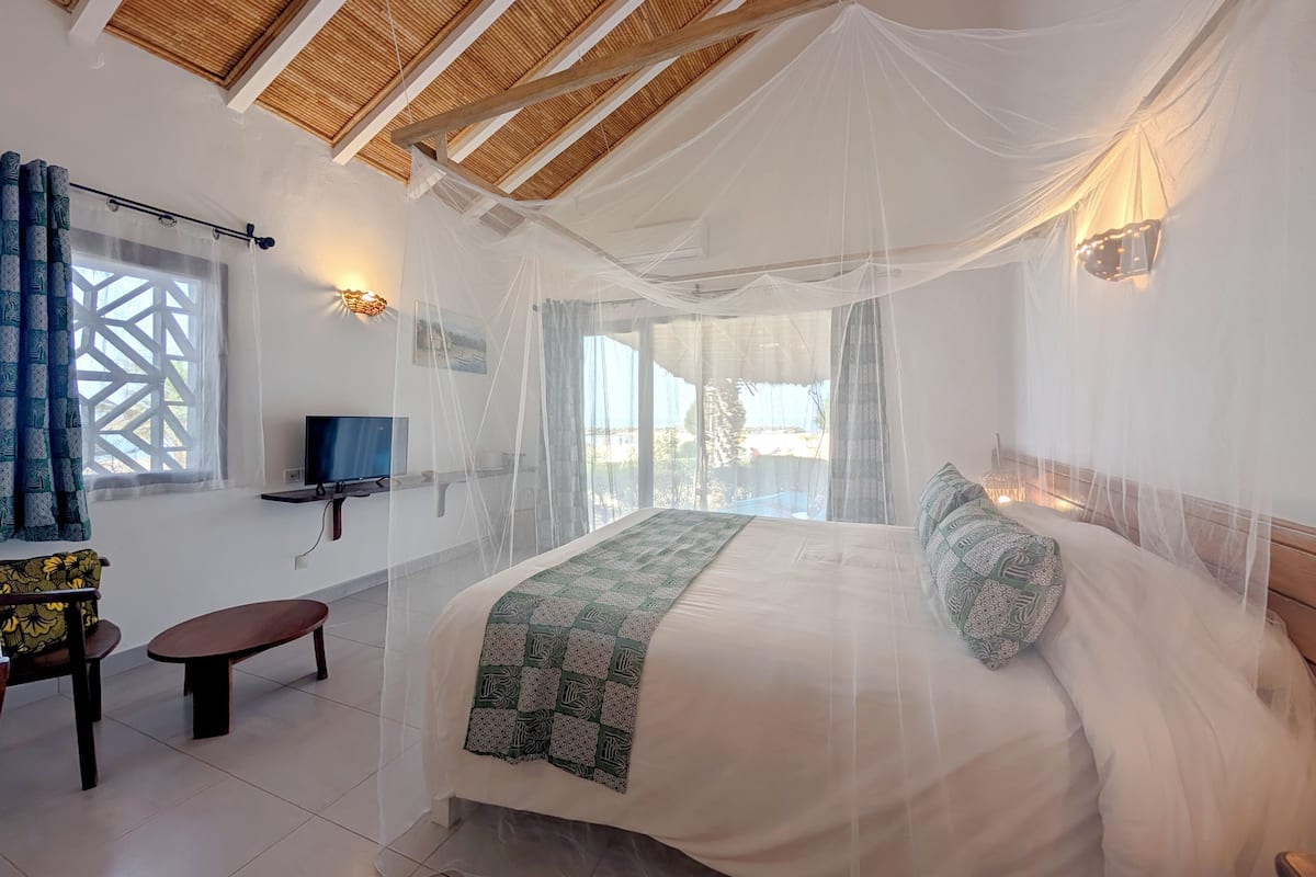 A bedroom with a bed, a TV, a bench, and a window with curtains.