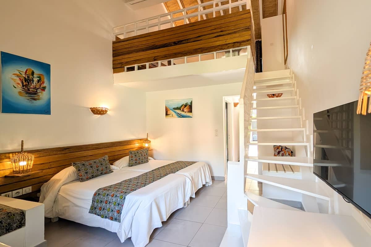 A hotel room with a wooden ceiling, a staircase, a bed with patterned bedding, a flat-screen TV, and artwork on the walls.