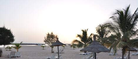 A beach with white lounge chairs and thatched umbrellas, palm trees, and a clear sky.