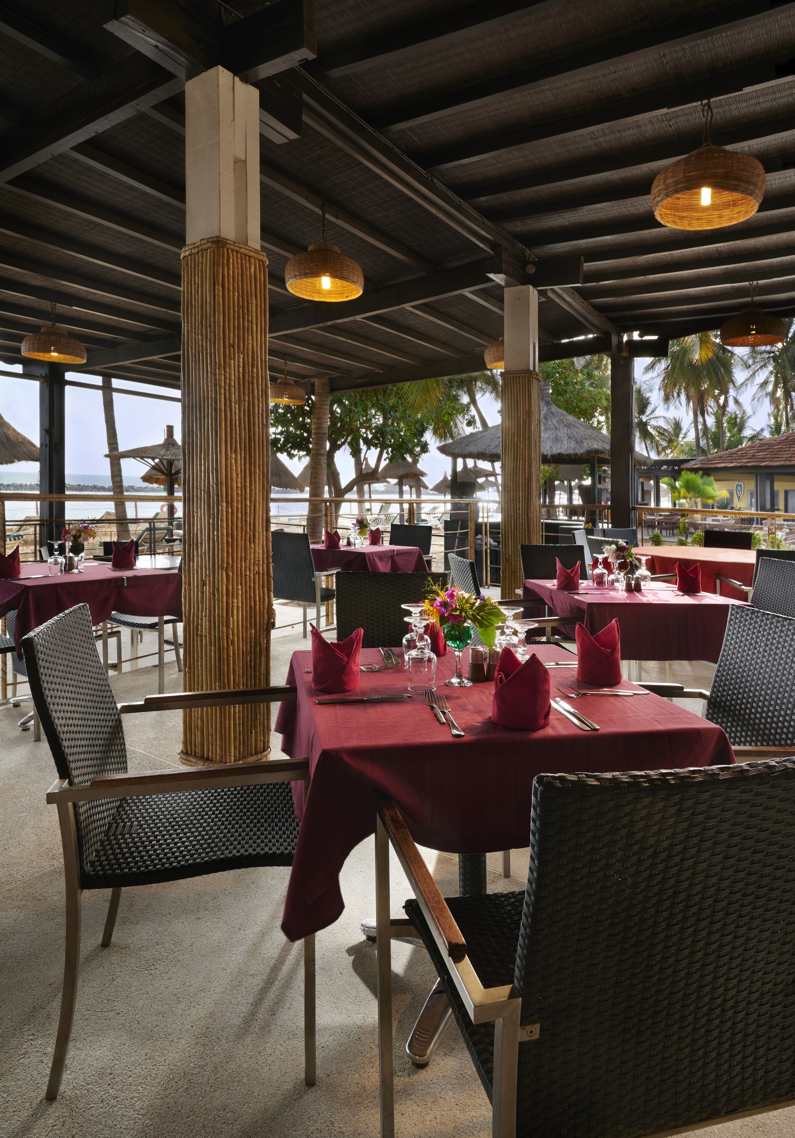 An outdoor restaurant with tables set for dining, featuring wicker chairs and red tablecloths.