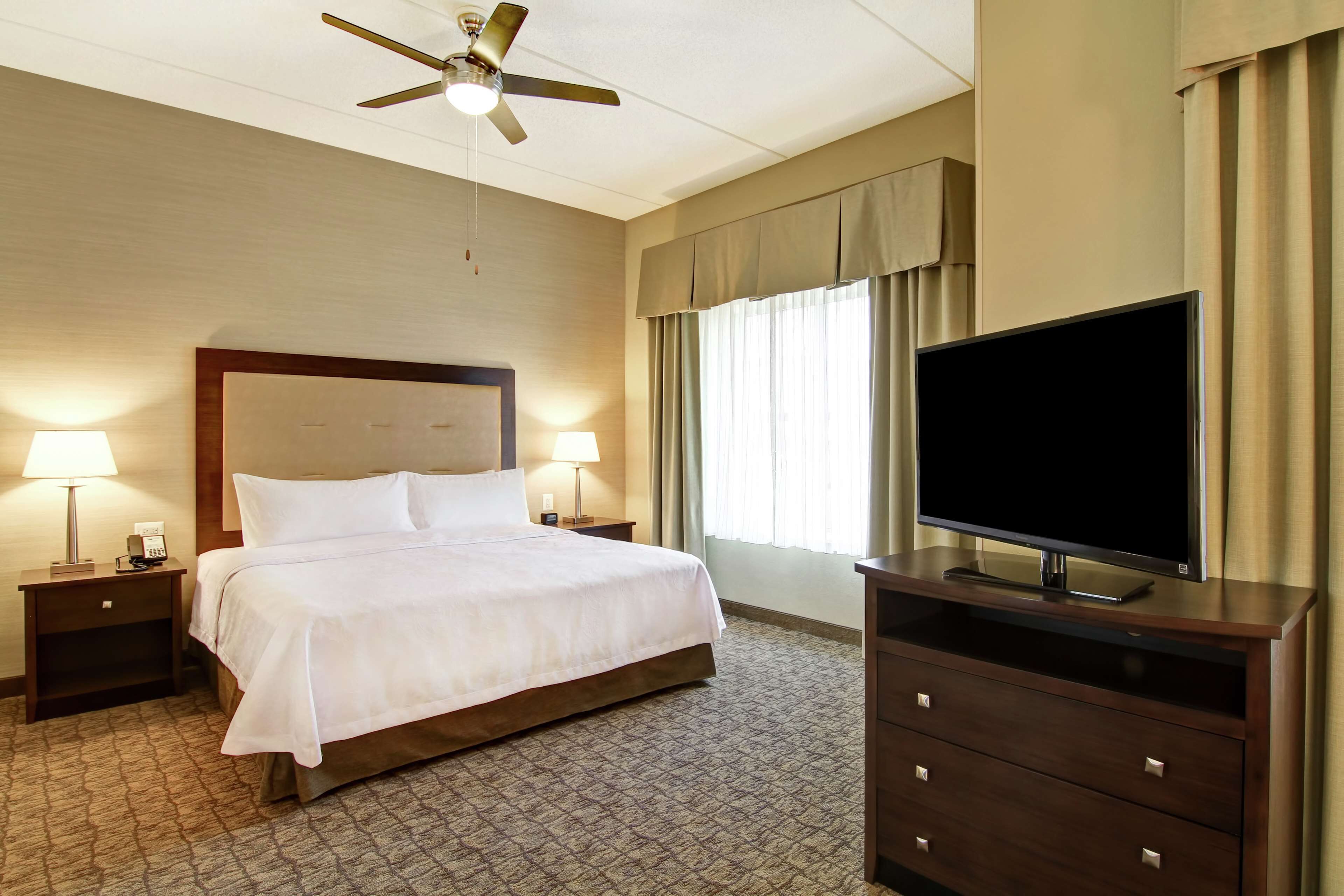 Una habitación de hotel con una cama, dos mesitas de noche con lámparas, un televisor en un aparador, un ventilador de techo y una ventana con cortinas.