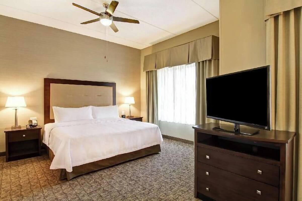 Une chambre d’hôtel comprenant un lit, deux tables de chevet avec des lampes, un ventilateur de plafond, un téléviseur à écran plat posé sur une commode, et une grande fenêtre avec des rideaux.