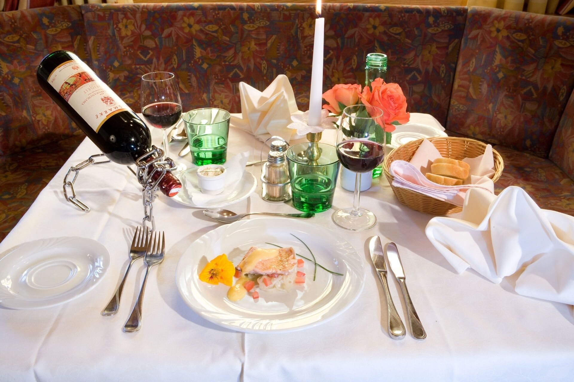 Una mesa de comedor dispuesta con una botella de vino, copas, cubiertos y un plato de comida.