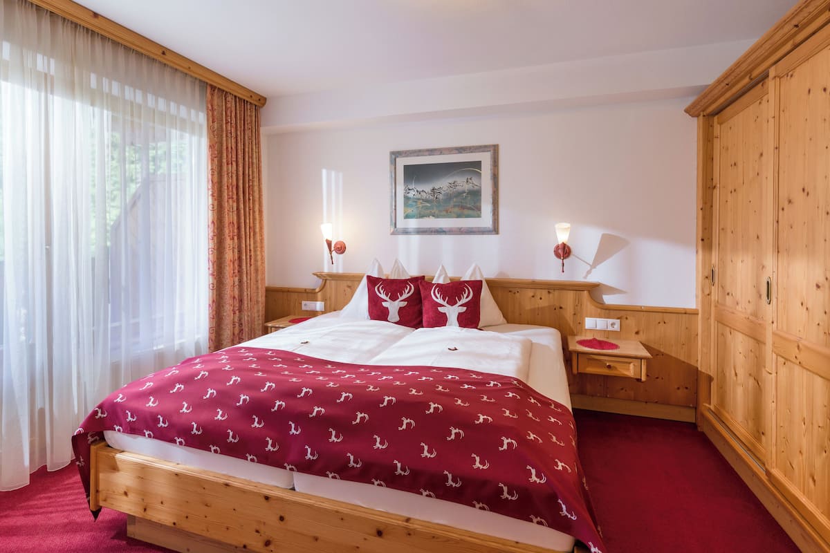 A hotel room with a wooden bed, a large window with sheer curtains, a wooden wardrobe, and a framed picture on the wall.
