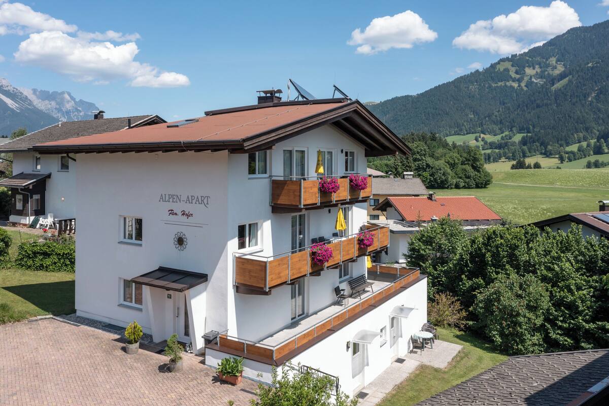 A two-story building with a sign reading "ALPEN-APART". It features a balcony with wooden railings and potted plants.