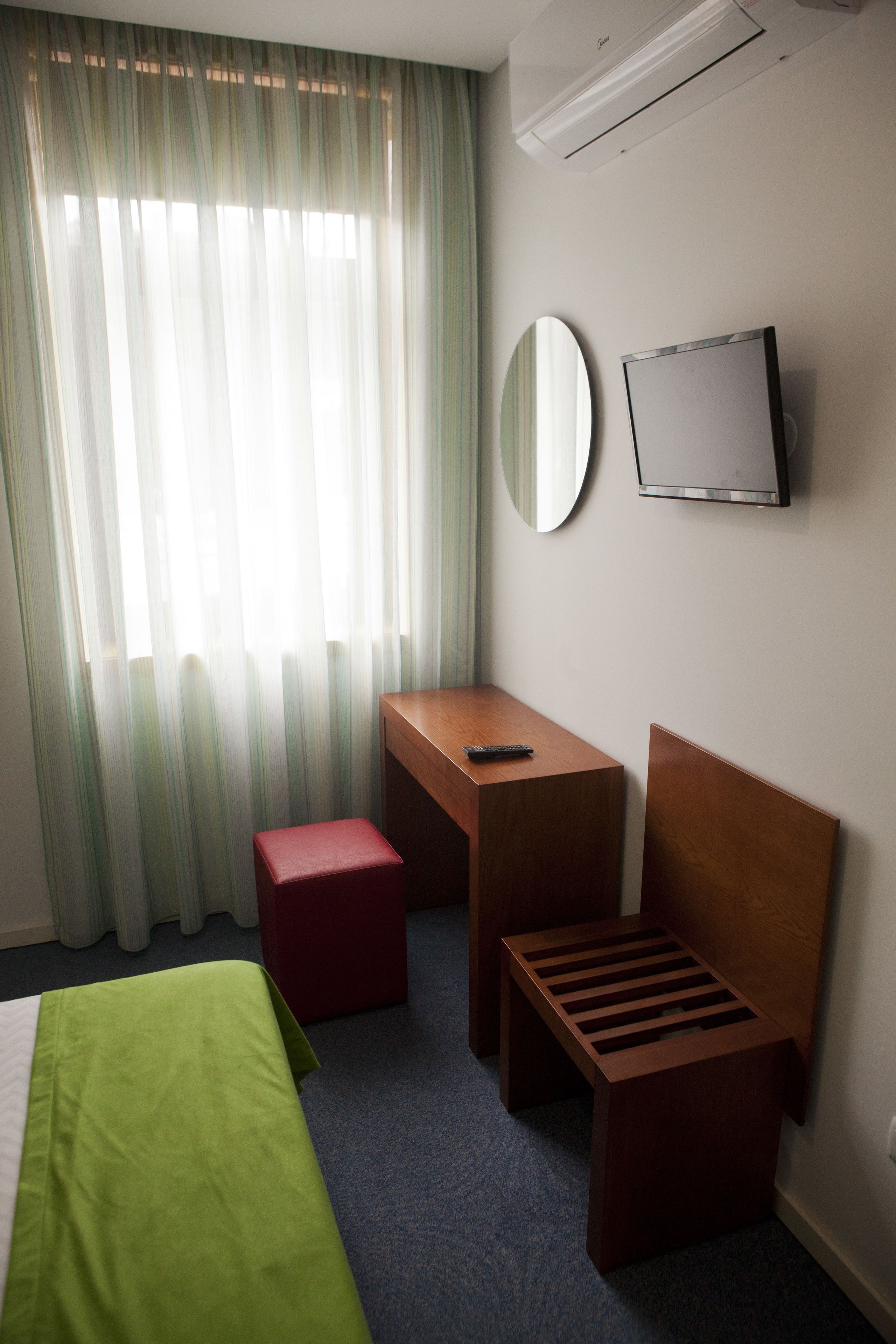 A hotel room with a bed, a desk, a chair, a small stool, a mirror, and a television.