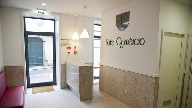 Zona de recepción de un hotel con un mostrador, un banco rojo y un cartel en la pared que dice 'Hotel Comercio'.