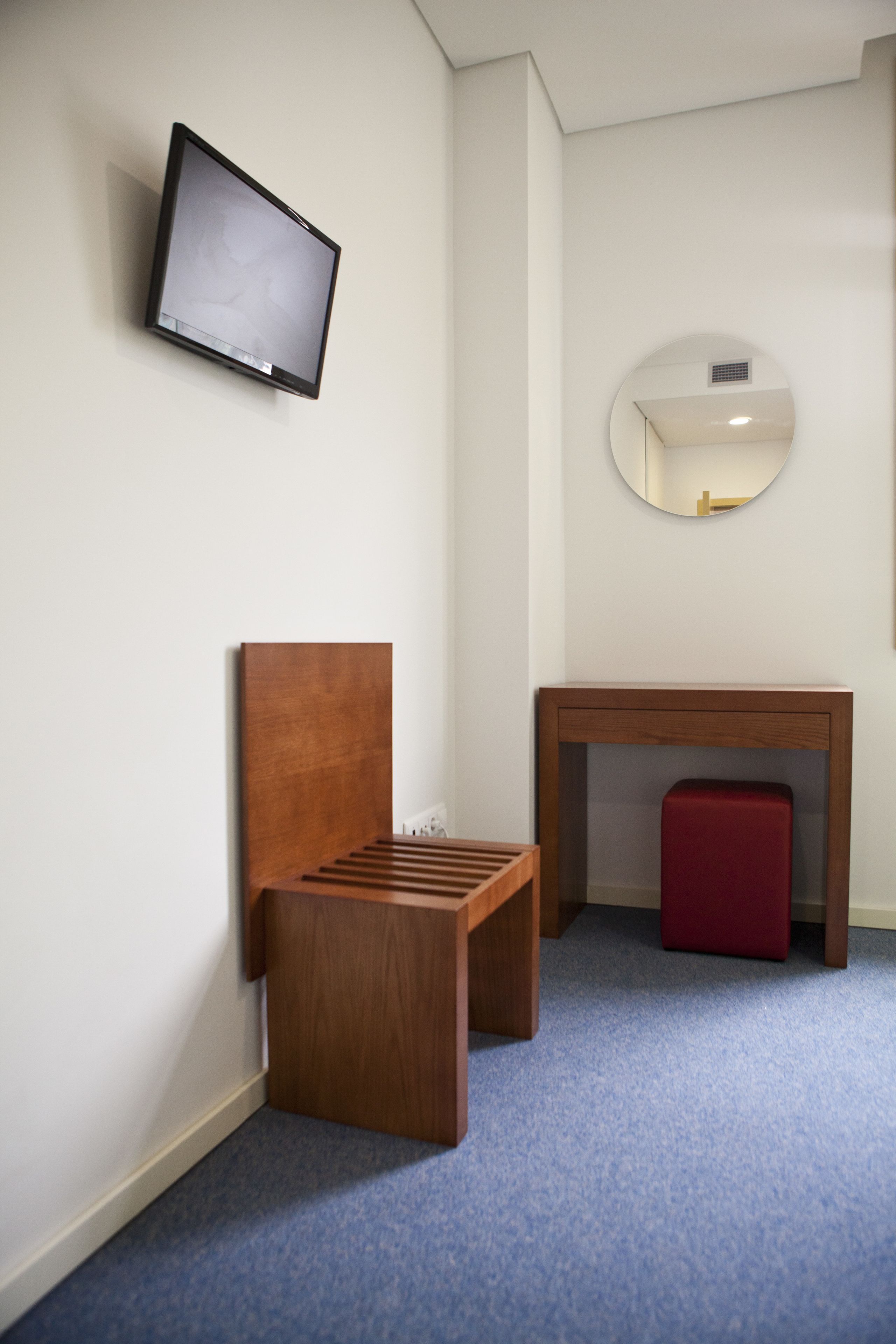 Een kamer met een houten bankje, een kleine tafel, een rode kruk, een aan de muur bevestigde televisie en een spiegel.