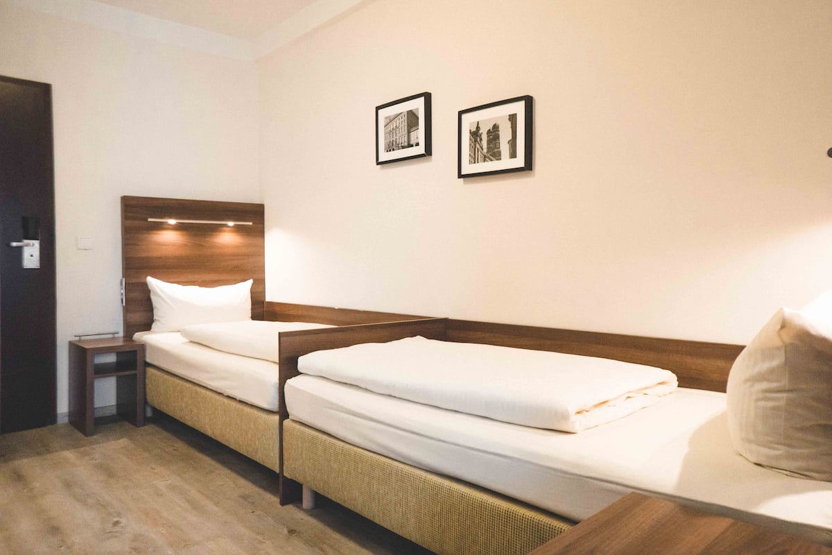 Una habitación de hotel con dos camas individuales, una mesita de noche pequeña y dos cuadros en la pared.