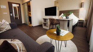 A modern living room with a sofa, a coffee table, a TV, and a dining area with a chair and a plant. - Hotel Säntis (Munich)