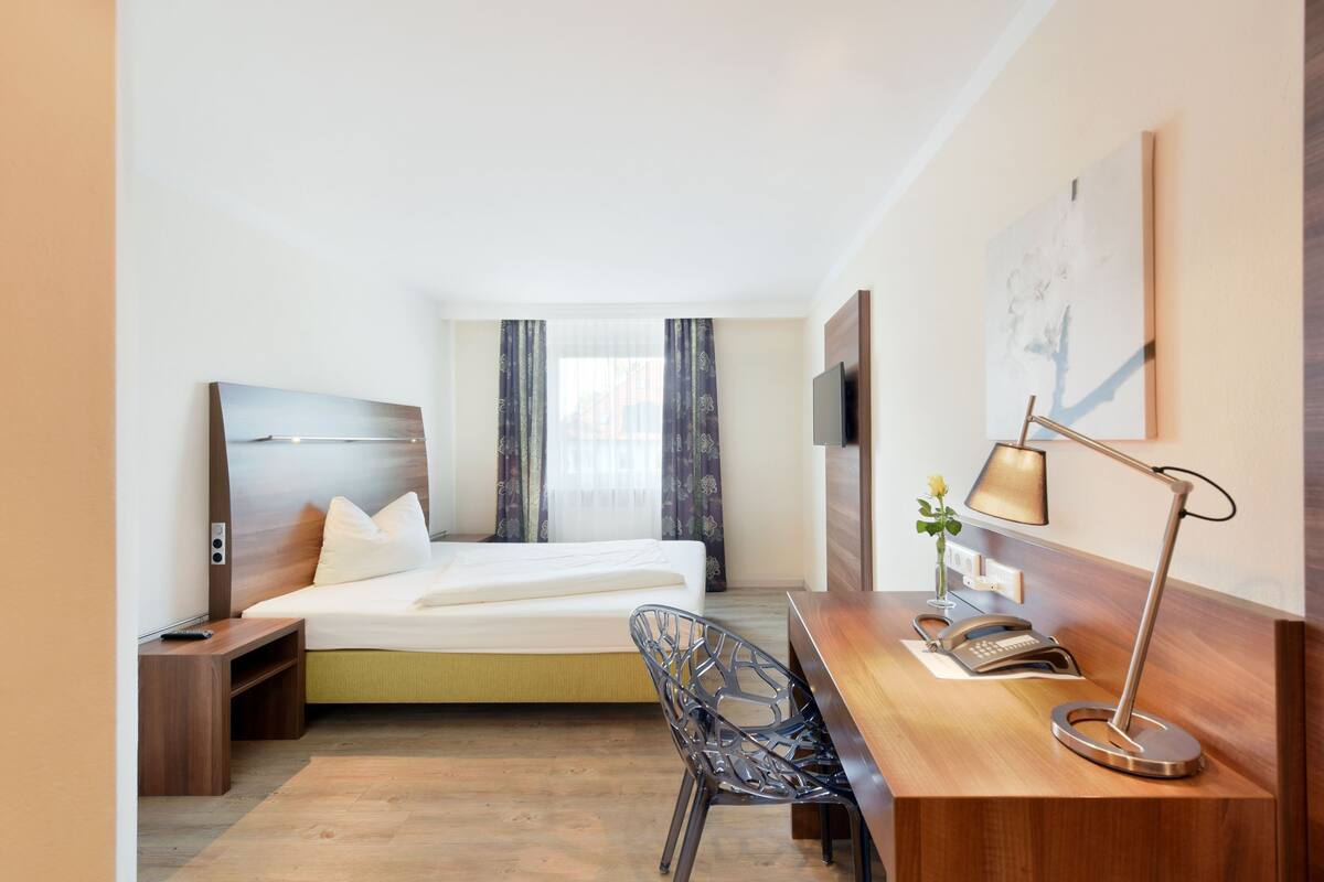 Habitación de hotel con cama, escritorio con lámpara, una silla, televisor y un cuadro en la pared.