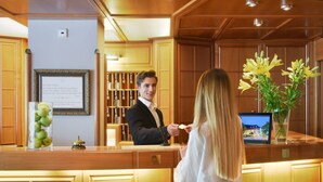 A reception desk with a framed sign, a vase of yellow flowers, and a digital display. - Hotel Säntis (Munich)
