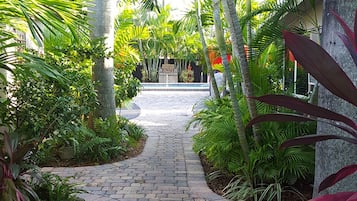A paved pathway flanked by lush tropical plants and palm trees, leading to a building with a stone facade.