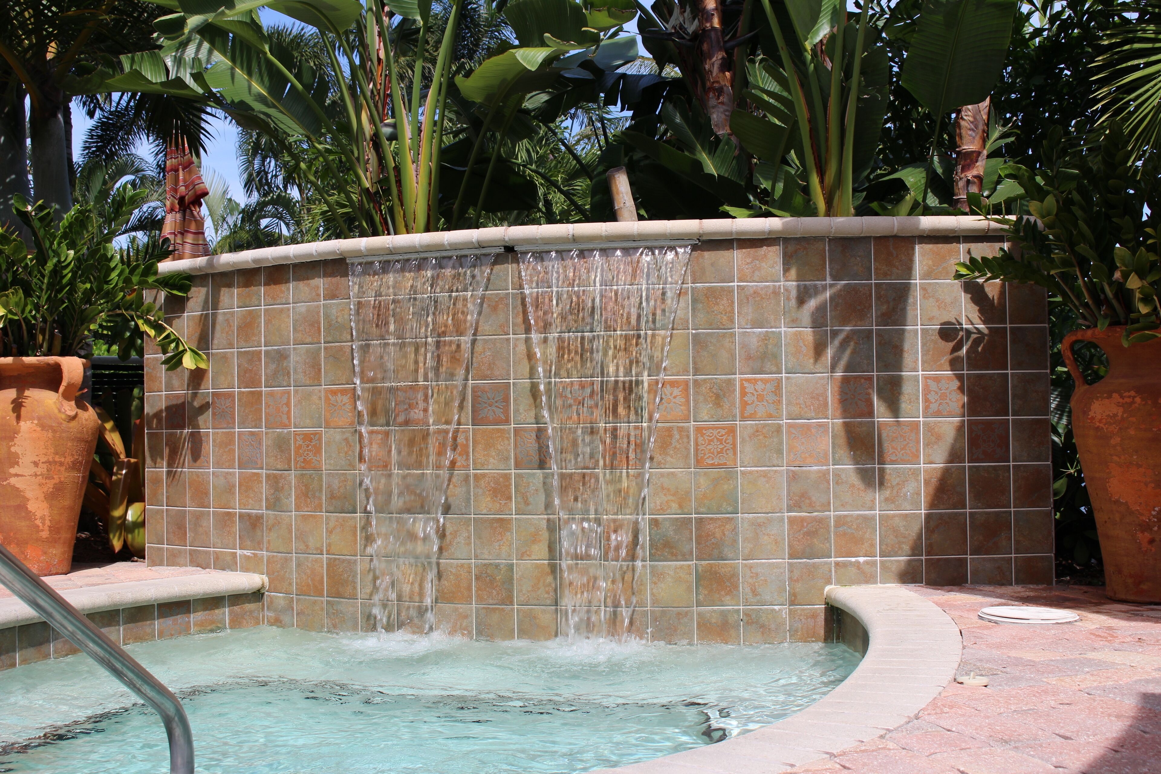 A pool with a tiled waterfall feature, surrounded by potted plants and a tiled wall.