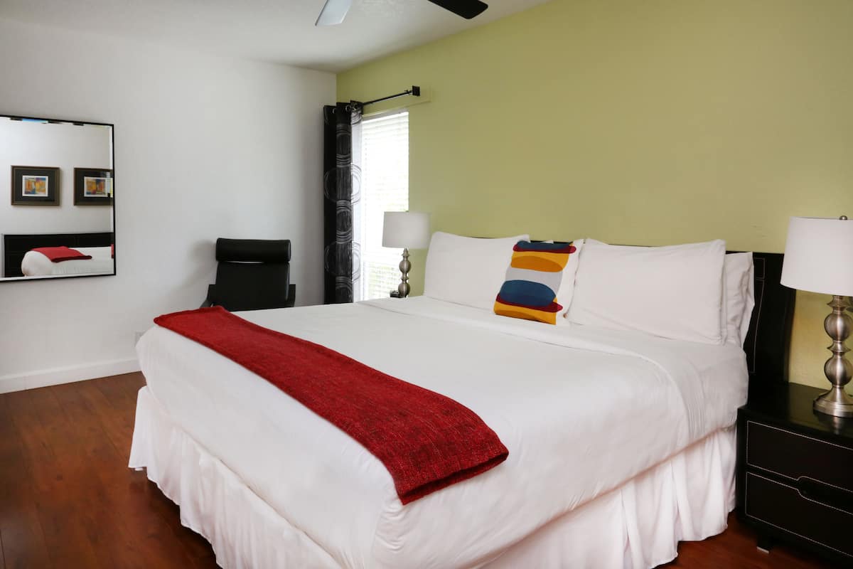 Una camera d'albergo con un letto grande, una coperta rossa, un comodino con una lampada e uno specchio a parete.