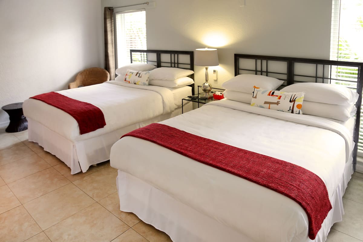 Camera d'albergo con due letti, ognuno con biancheria da letto bianca e una coperta rossa. C'è un comodino con una lampada e un tavolino con una ciotola rossa.