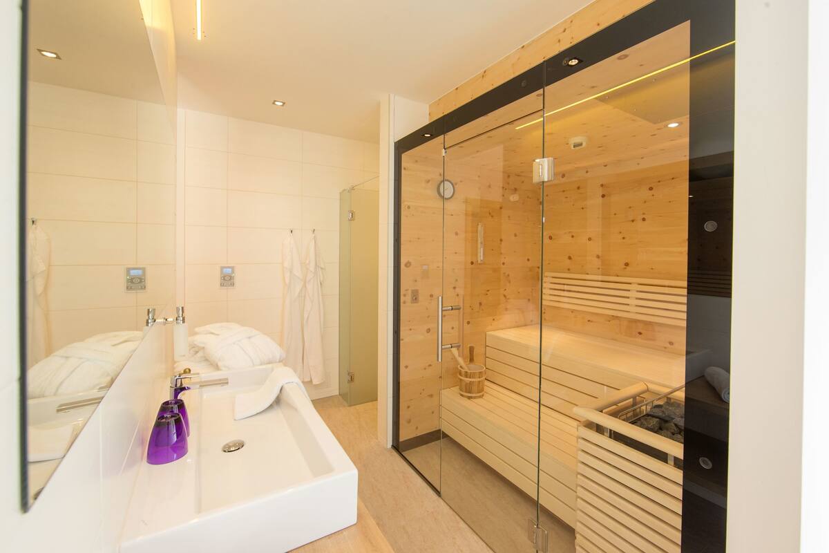 A modern bathroom with a glass-enclosed sauna, a white sink with a purple soap dispenser, and a large mirror.