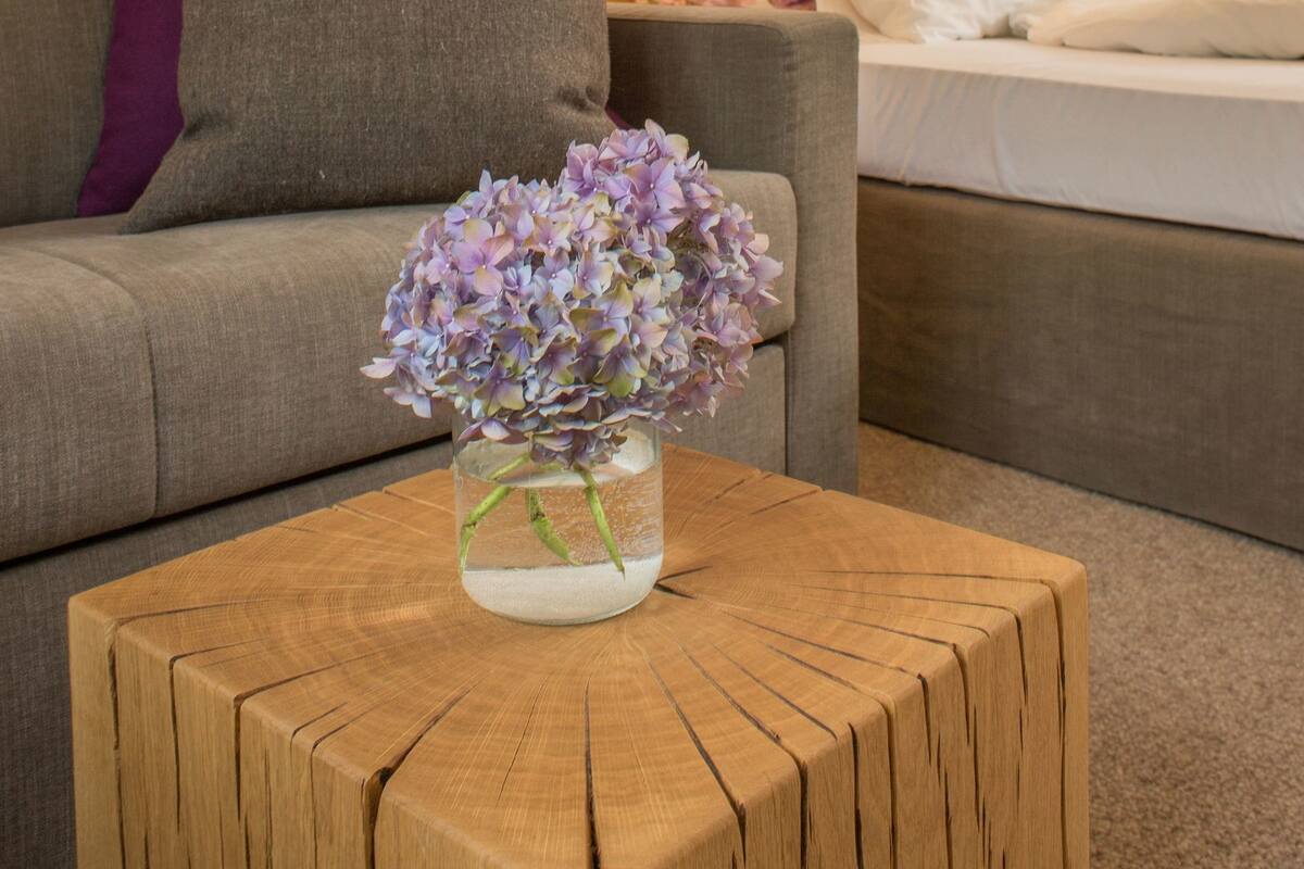 A cozy room with a wooden wall, a grey sofa with a purple pillow, a wooden coffee table with a vase of flowers, and a bed with white bedding and floral pillows.