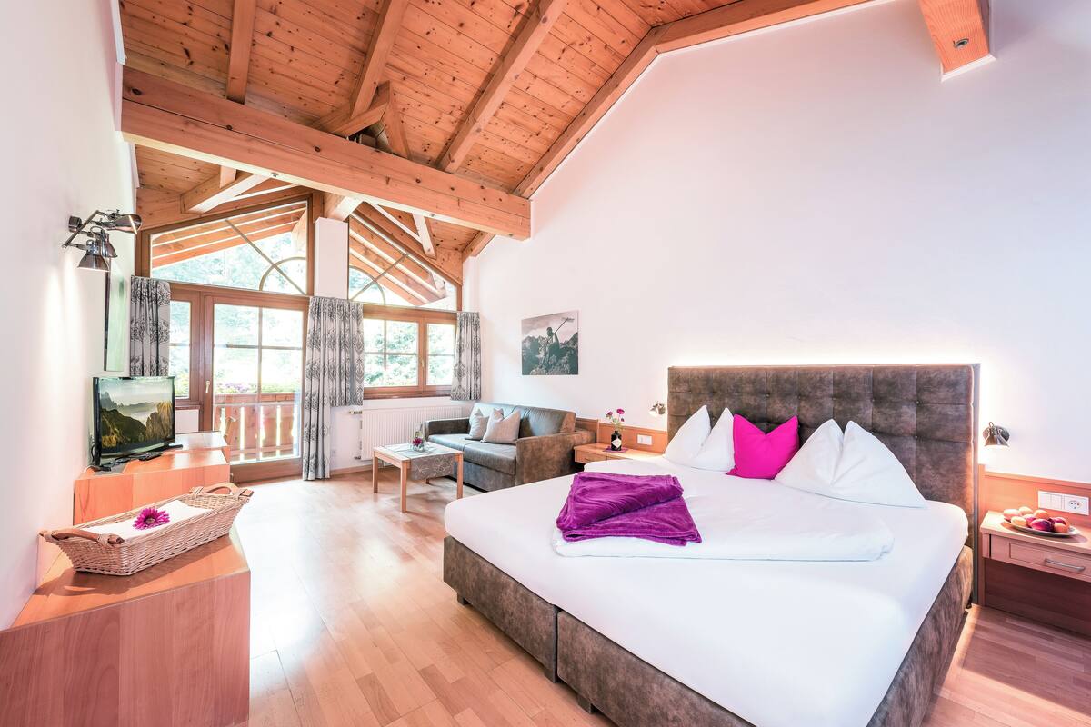 A spacious bedroom with a large bed, wooden ceiling beams, a wooden desk, and a view of the outdoors through a large window.