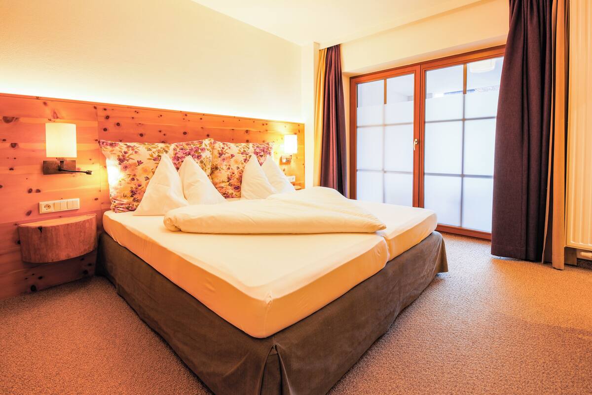 A hotel room with a wooden headboard, a large bed with white bedding, a nightstand with a lamp, and a view of the outside through a sliding glass door.