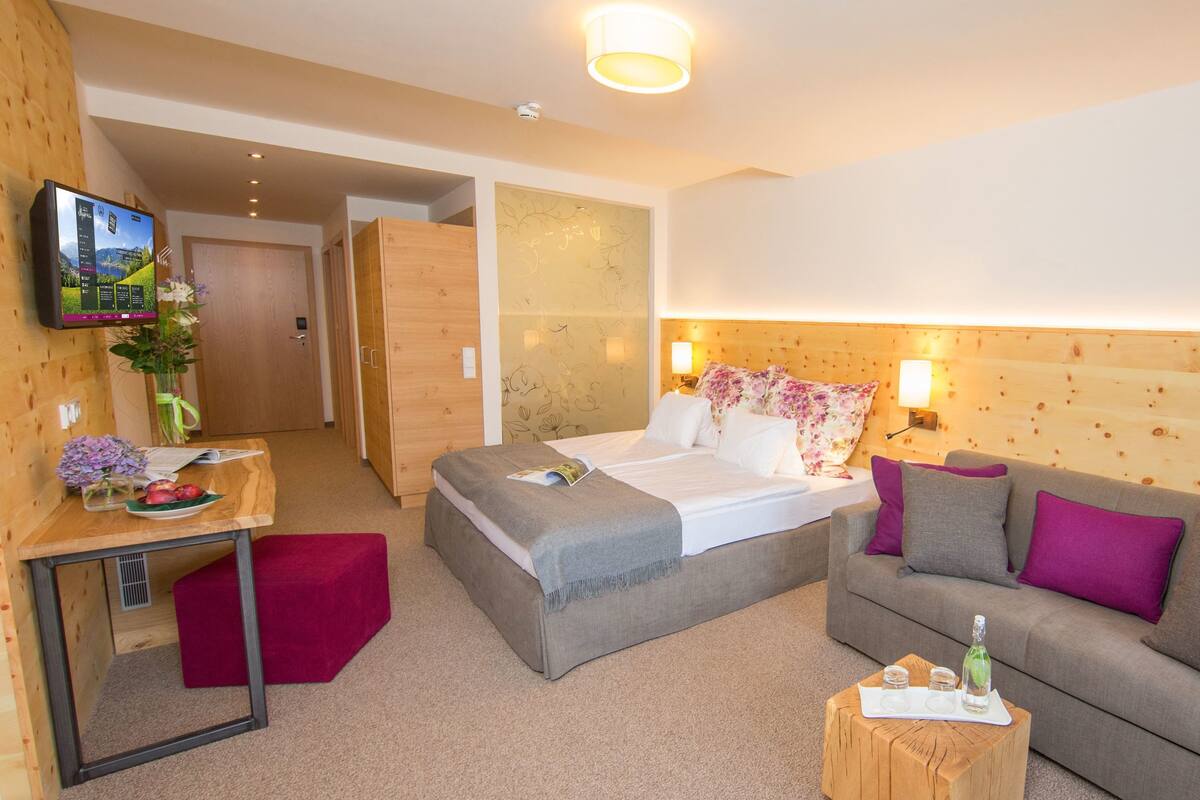 A hotel room with a bed, a sofa, a small table with a bottle and glasses, a TV on the wall, and a wooden door.