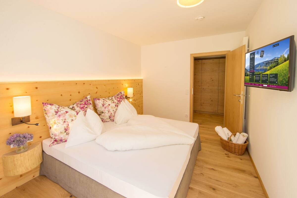 A hotel room with a wooden headboard, a bed with white bedding and floral pillows, a bedside table with a lamp, a wall-mounted TV, and a basket of towels.