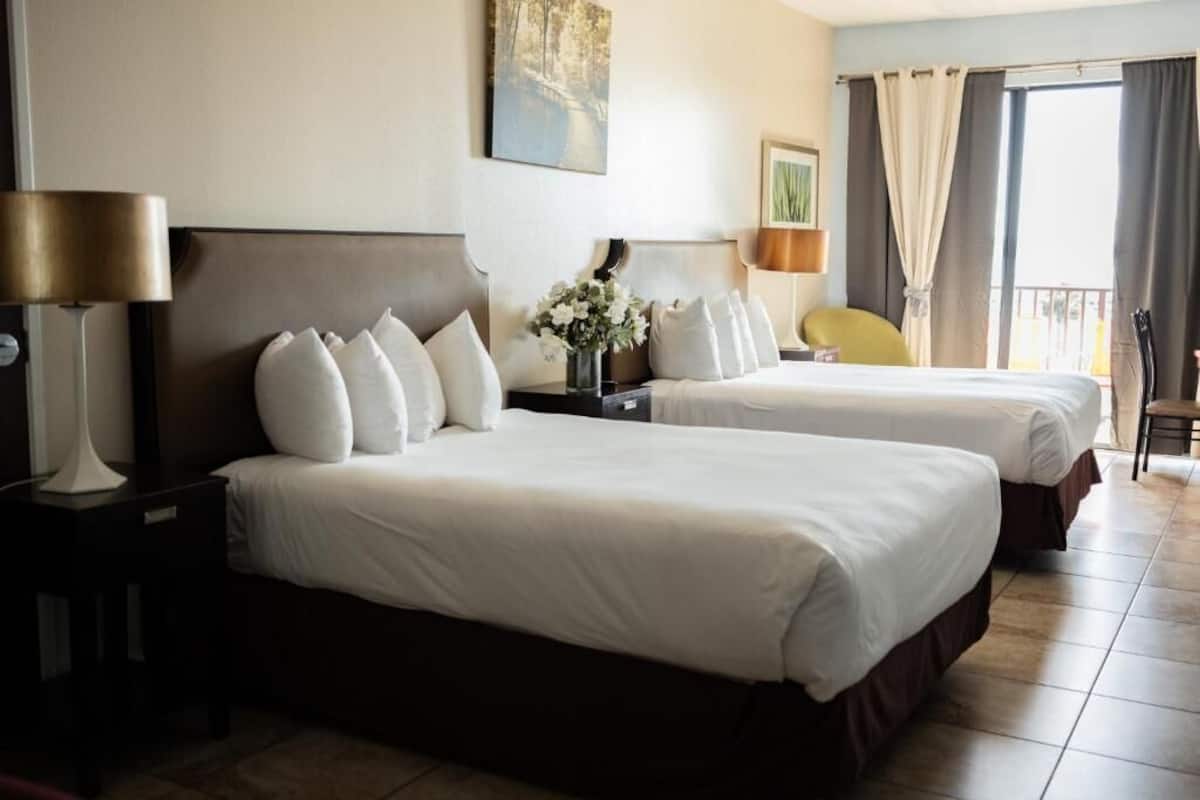 Ein Hotelzimmer mit zwei Betten, einem Nachttisch mit Lampe, einem Sessel und einem Balkon mit Vorhängen.