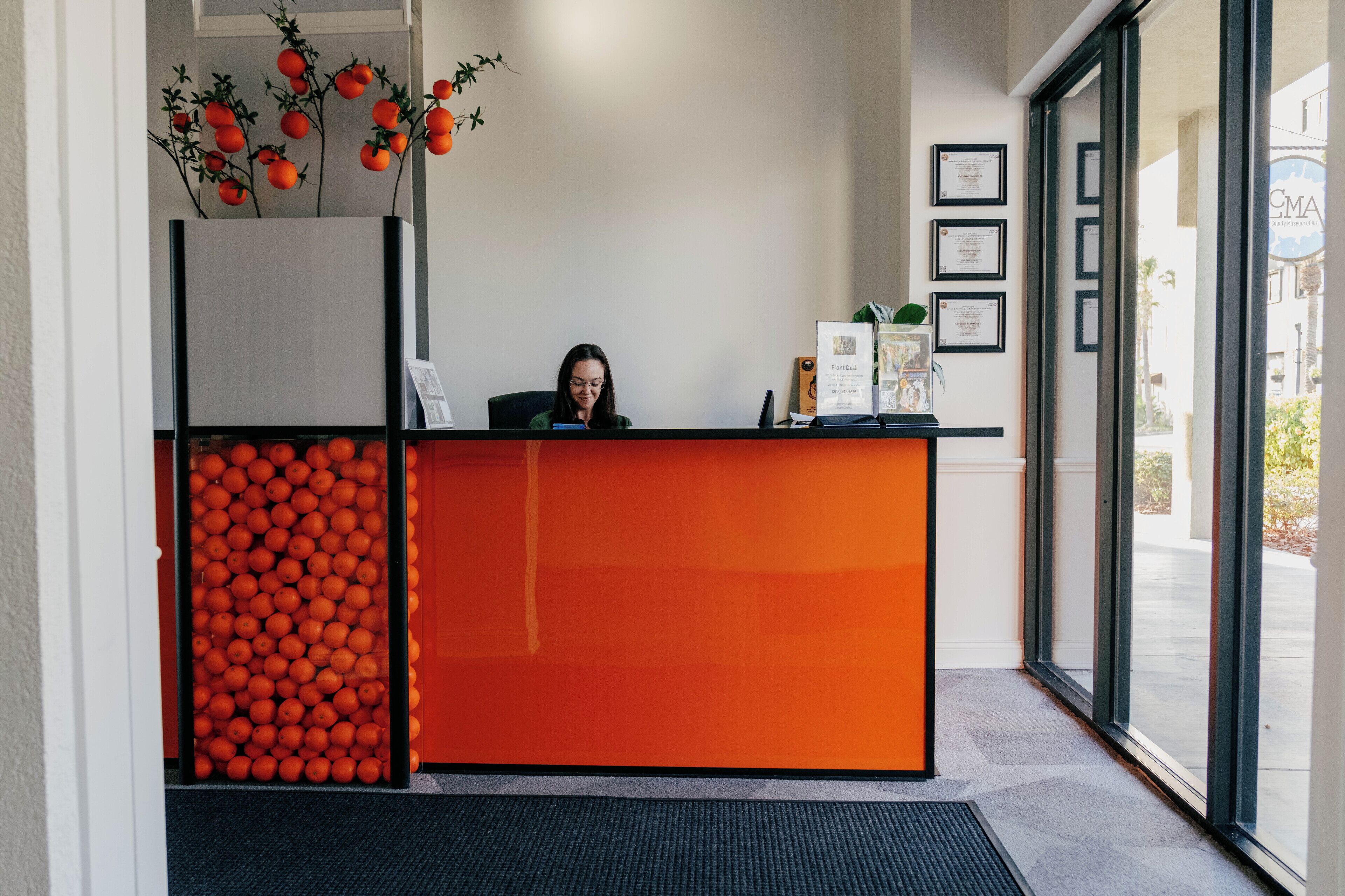 A reception desk with a woman seated behind it, a wall with framed certificates, and a large display of oranges.