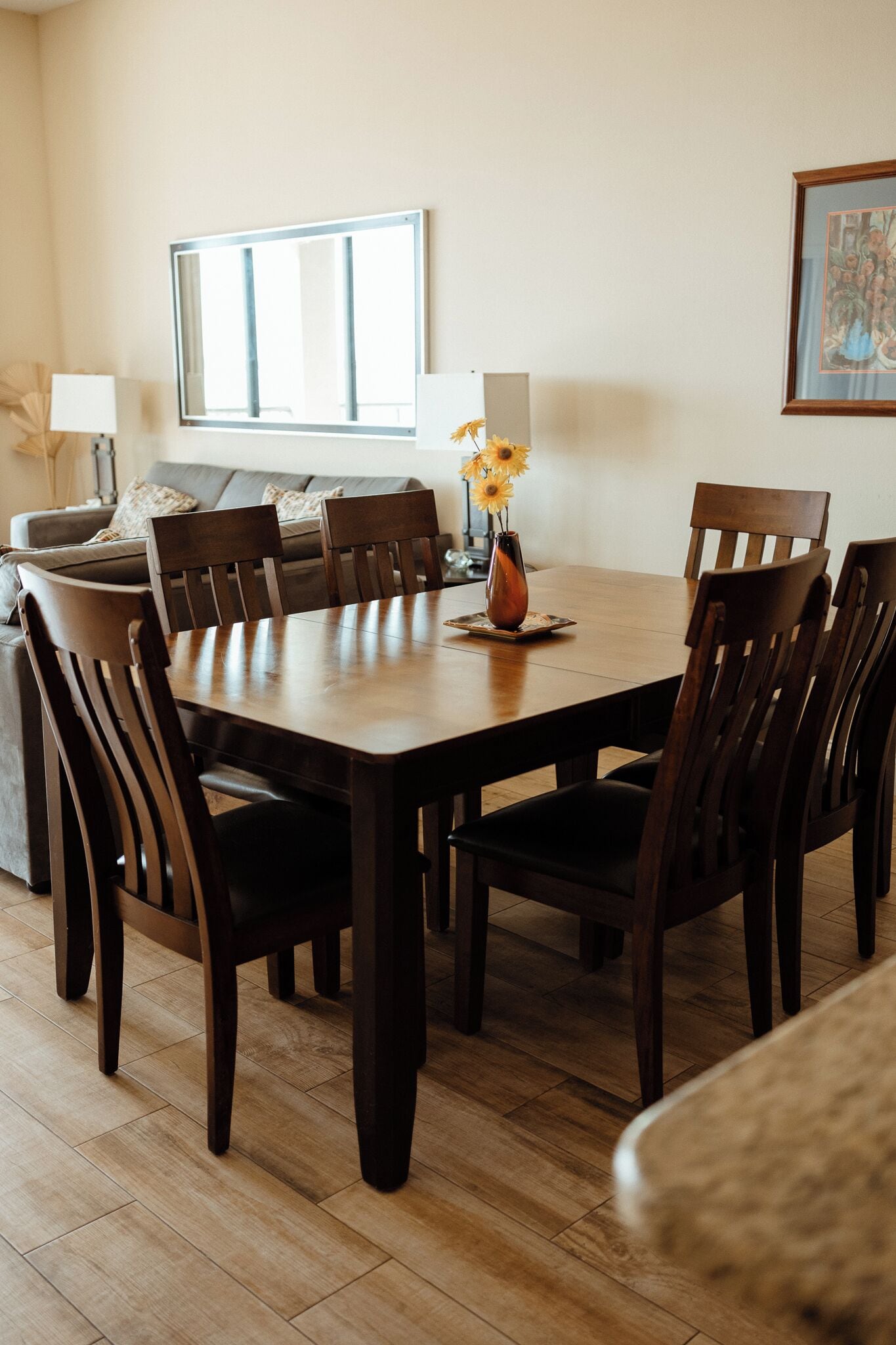 A dining area with a wooden table, chairs, a vase with flowers, a sofa, and a window.