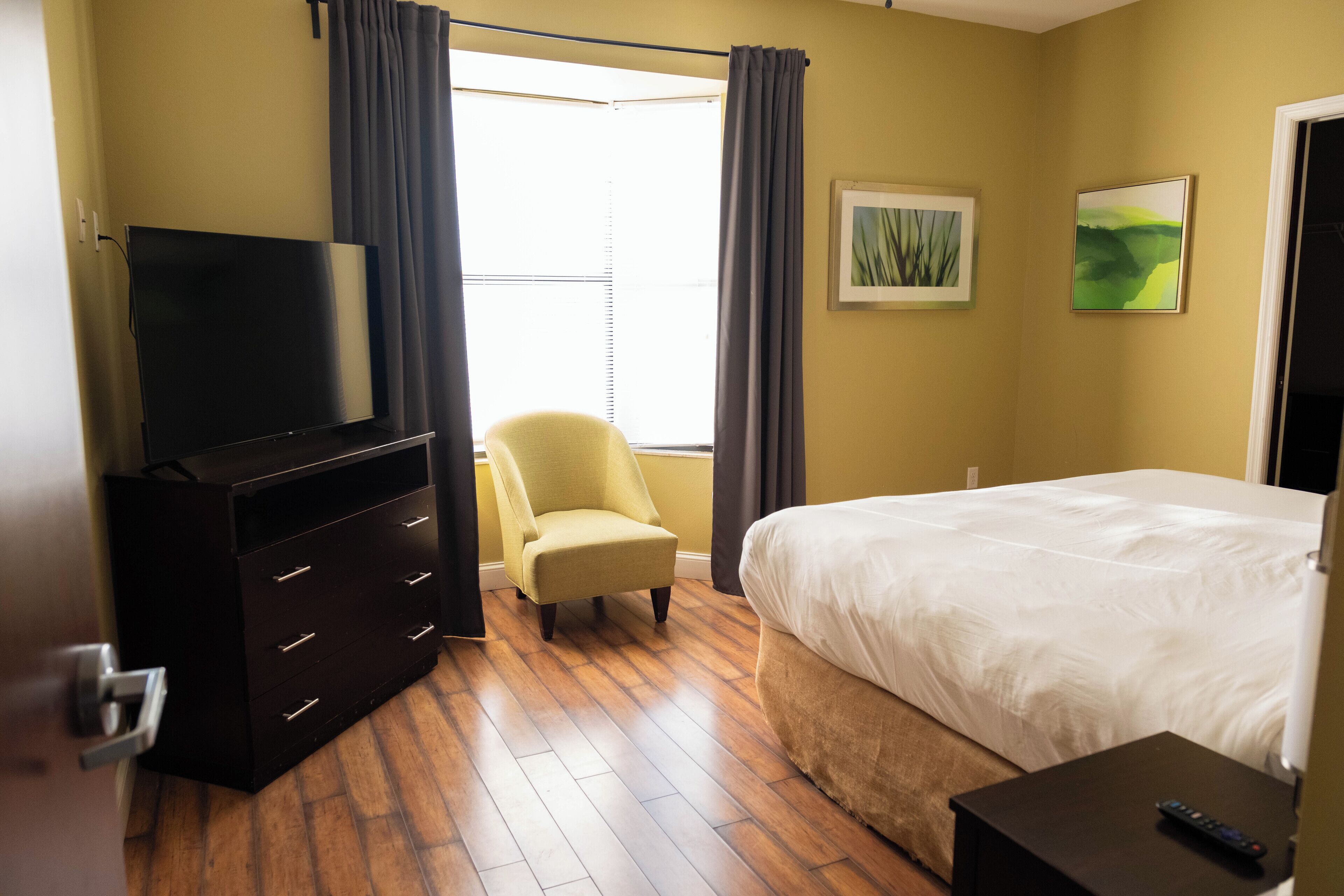 Een hotelkamer met een bed, een televisie op een dressoir, een stoel en een raam met gordijnen.