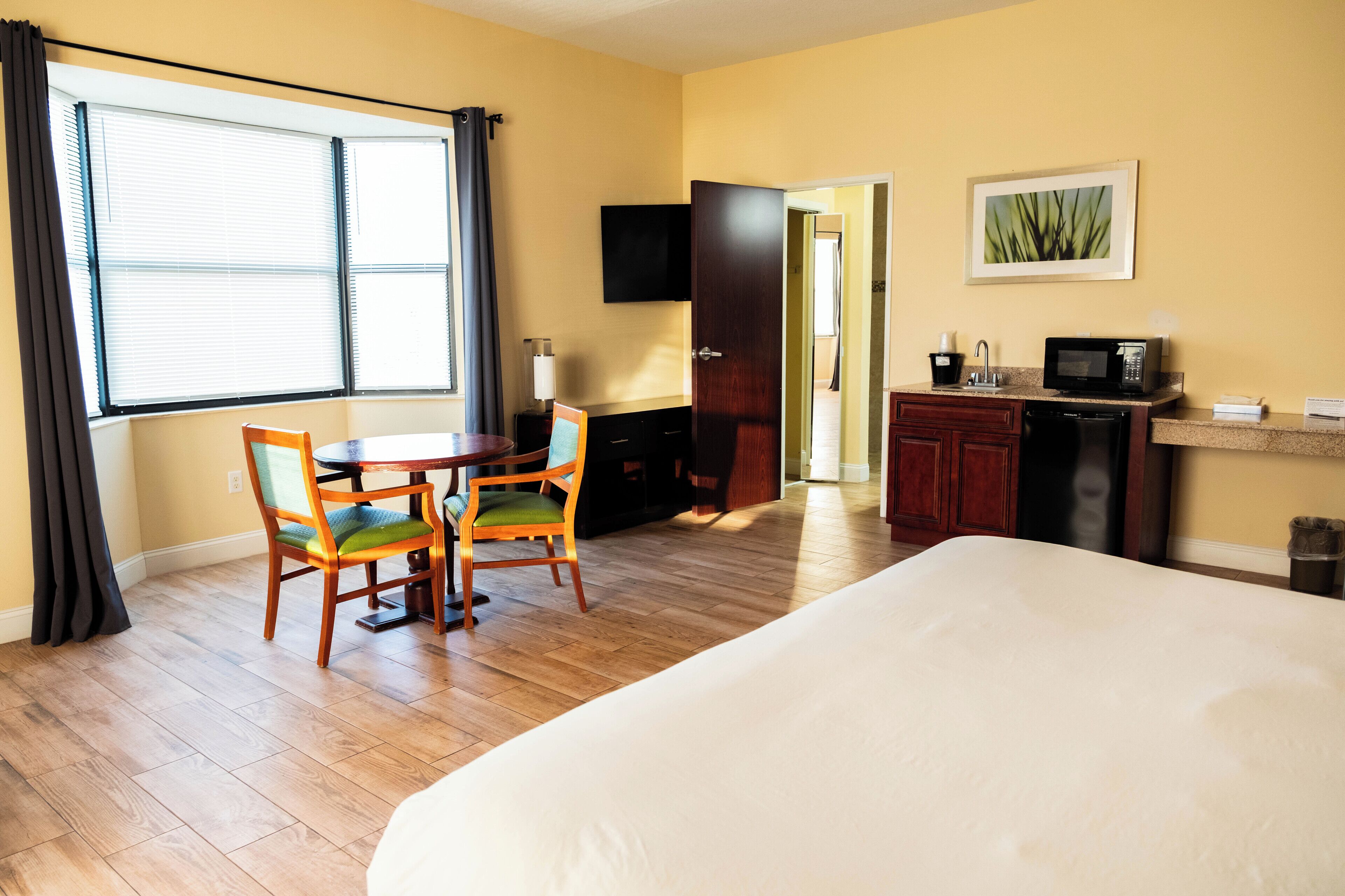 A hotel room with a bed, a dining table with chairs, a kitchenette, and a TV on the wall.