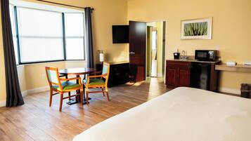 A hotel room with a bed, a dining table with chairs, a kitchenette, and a TV on the wall.