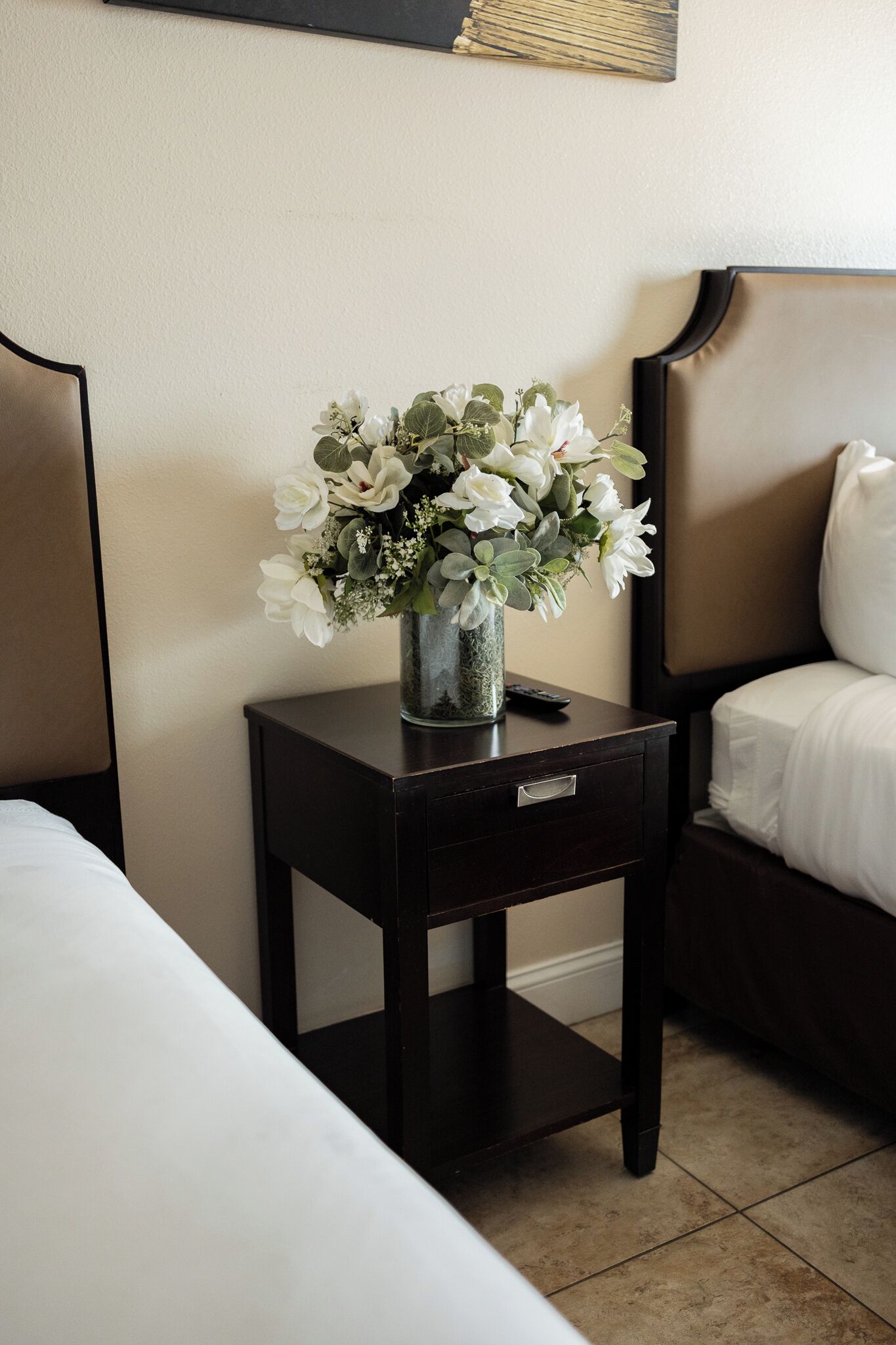 Habitación de hotel con una cama, una mesita de noche con un jarrón de flores blancas y un control remoto sobre la mesita.