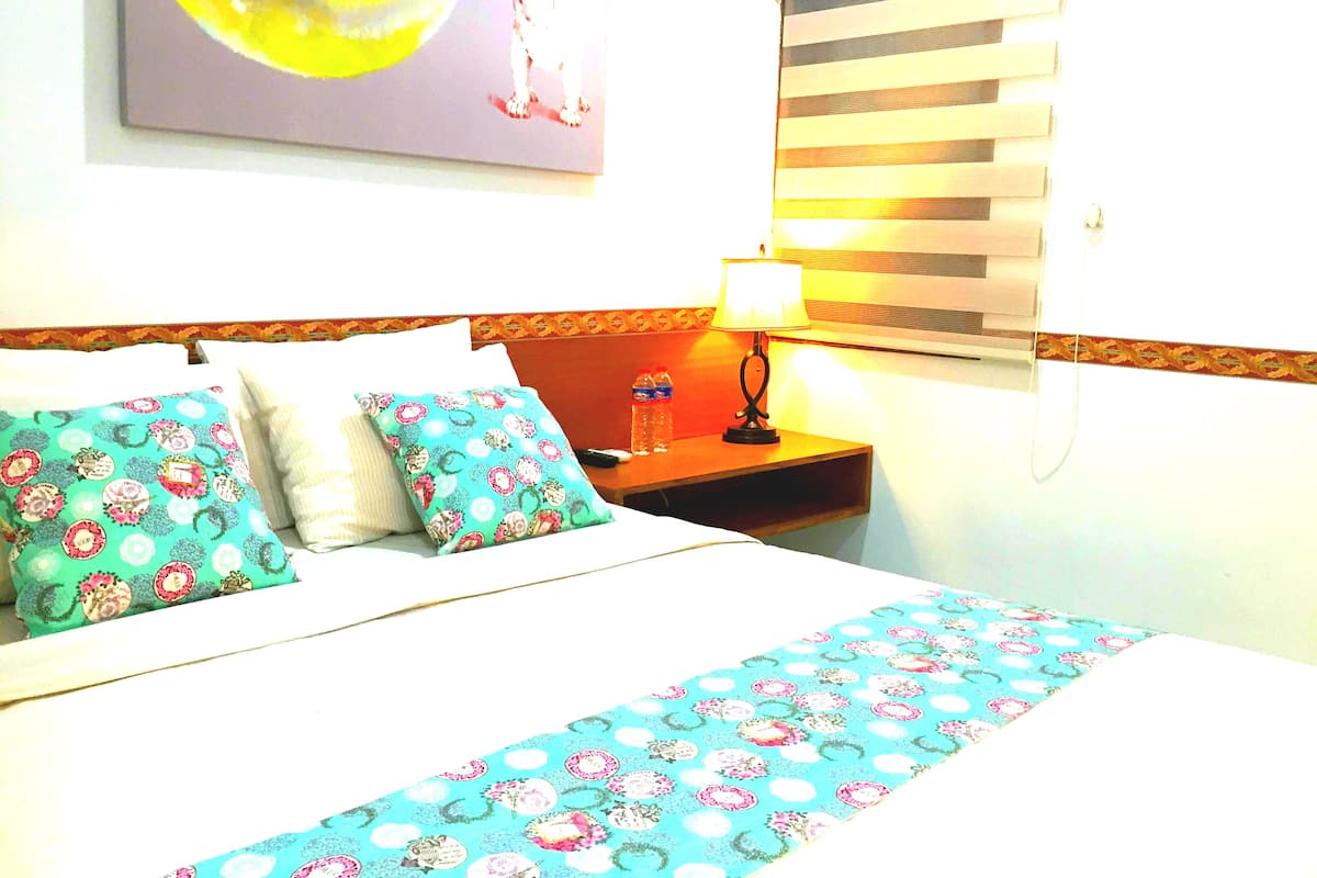 A hotel room with a bed, pillows, a nightstand, a lamp, a bottle, and a window with blinds.
