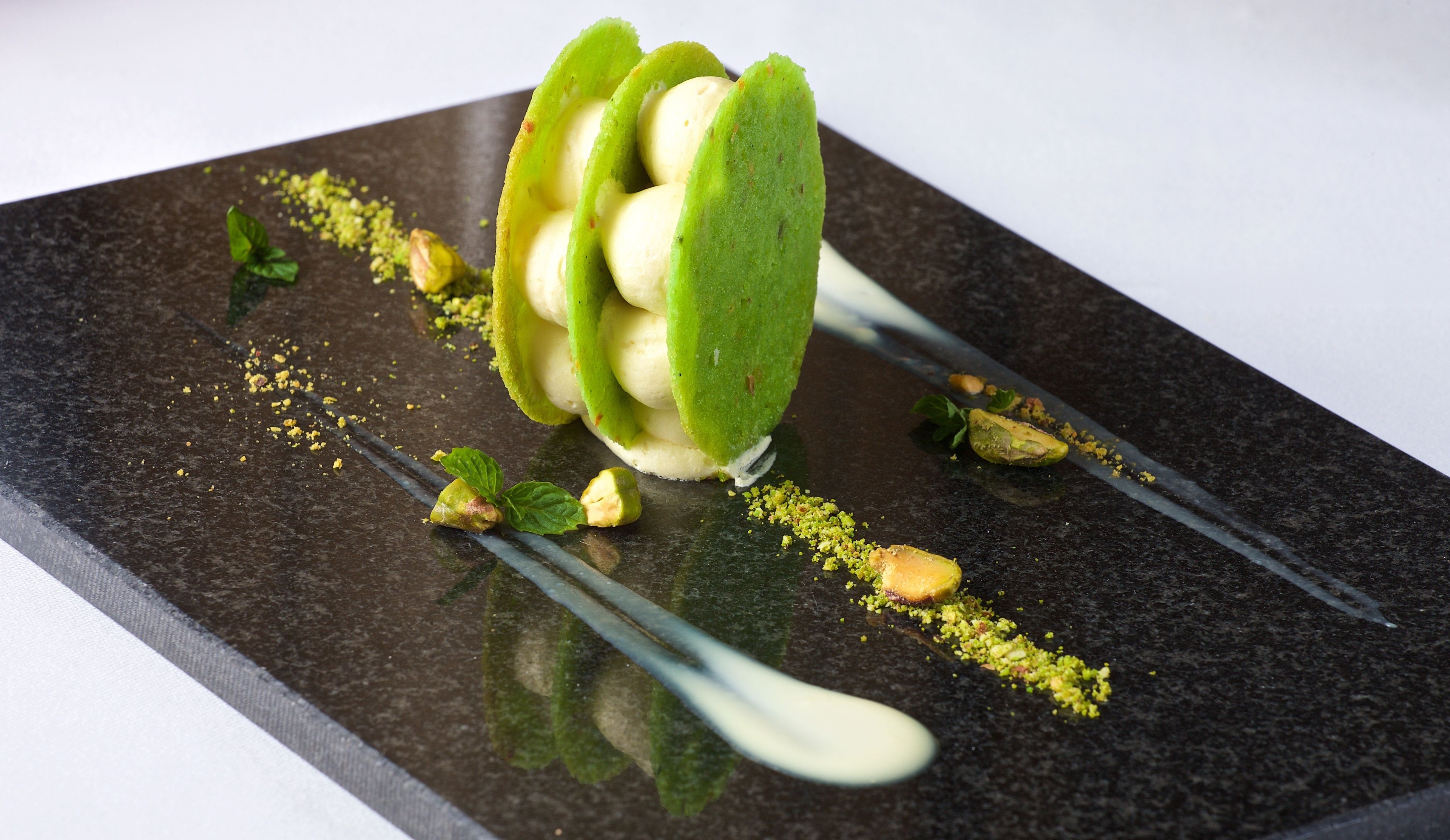 A dessert with green and white components, garnished with crushed pistachios and mint leaves, on a dark plate.