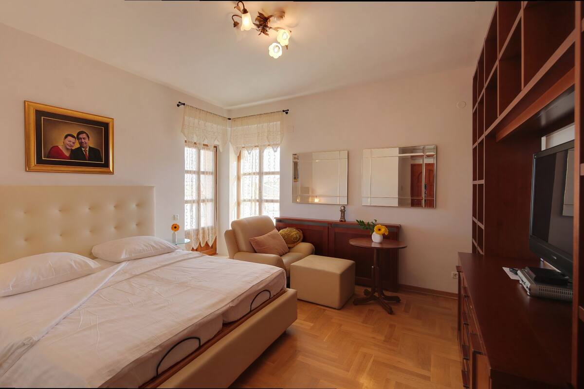 Un dormitorio con una cama grande, una mesita de noche con lámpara, una silla, un escritorio con espejo y un televisor.