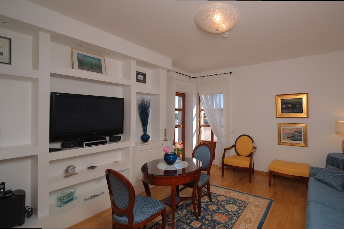 Una sala de estar con una televisión de pantalla plana, un sofá, una mesa de comedor con sillas y una mesita auxiliar con una lámpara.
