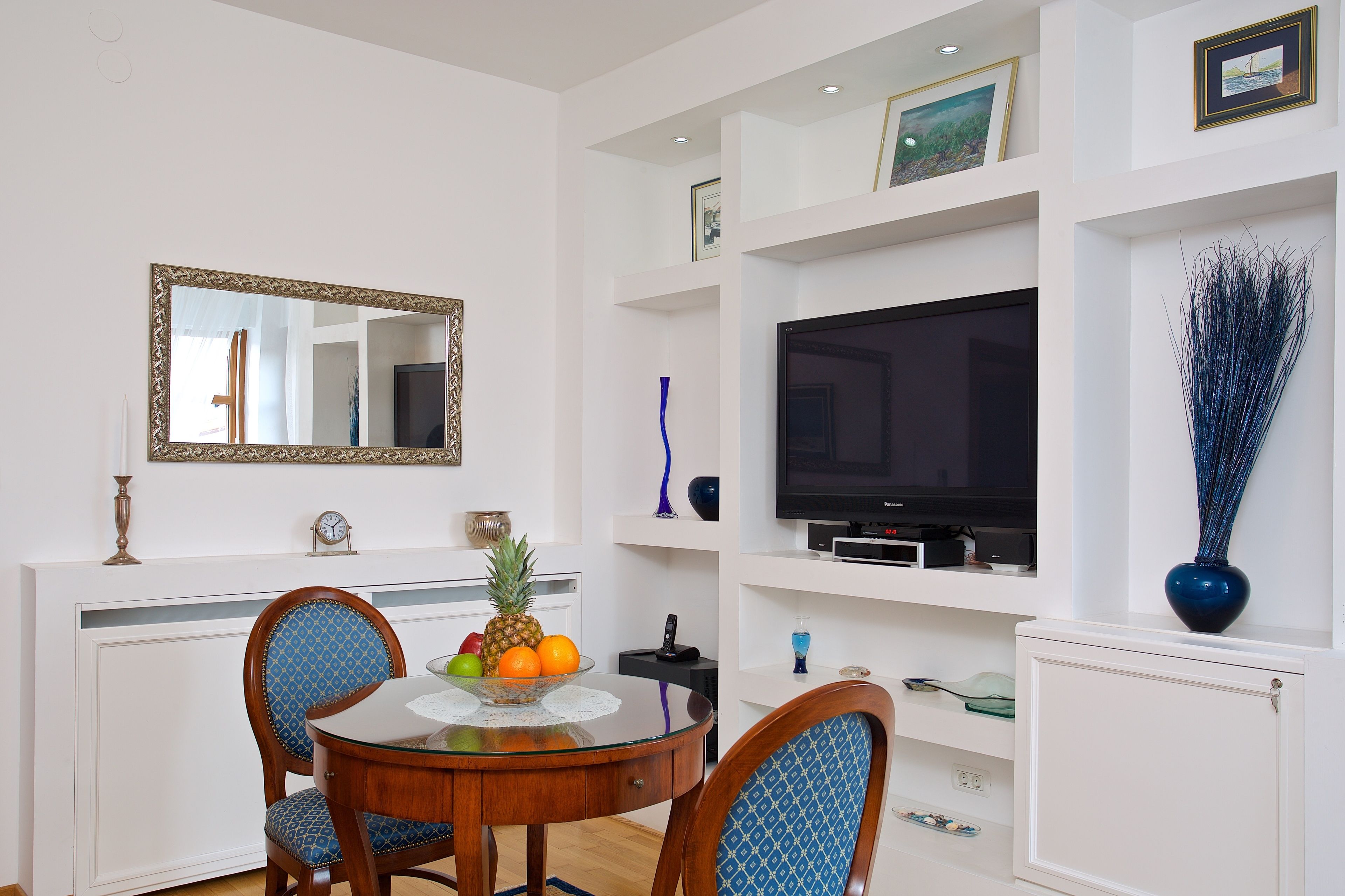 Un coin repas avec une table ronde, des chaises, un panier de fruits et un téléviseur fixé à un mur blanc.