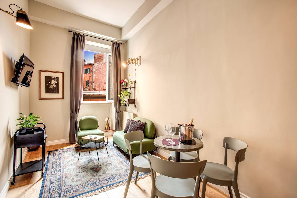 A cozy living area with a green sofa, a small round table with a wine glass and a bottle, and a plant on a side table.